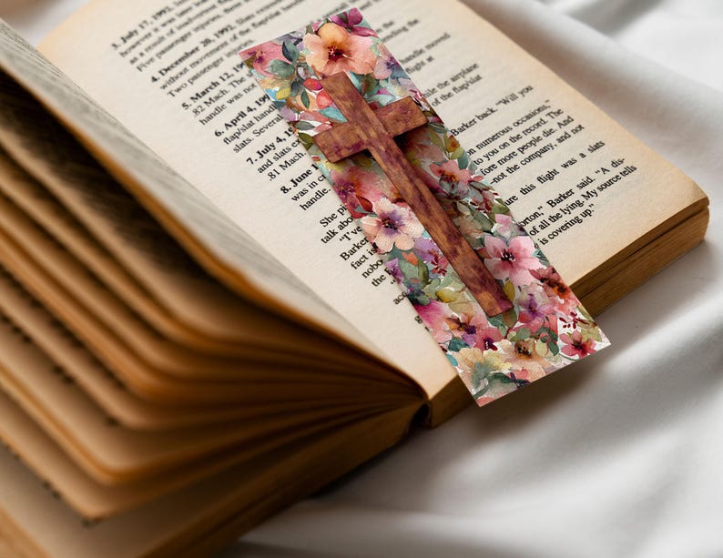 May include: A bookmark in the shape of a wooden cross, adorned with a floral pattern of pink, purple, and yellow blooms. It rests within an open book, its pages showing a vintage, aged appearance.