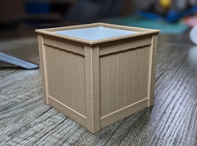 May include: A square, light brown planter with a white plastic liner. The planter has a wood-grain texture and is made of a durable material.