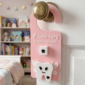 May include: A pink door hanger featuring a white tooth fairy icon, a small white box with a black tooth graphic, and the text "Tooth Fairy Please STOP Here". The sign is decorated with small white dots and stars, hanging from a brass-colored door knob.