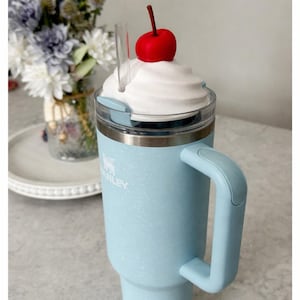 May include: A light blue Stanley tumbler with a handle, topped with a white whipped cream design and a red cherry. The tumbler has a clear straw and a silver rim. The tumbler is designed to keep drinks cold or hot.