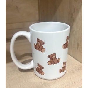 May include: White ceramic mug featuring a repeating pattern of brown teddy bear illustrations. The mug has a curved handle and a smooth, glossy finish. The teddy bears are depicted in a seated position.
