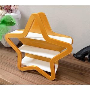 May include: A star-shaped, three-tiered shelf in a warm yellow-orange hue. The shelves are white, creating a contrast with the frame. The design is modern and geometric, suitable for displaying small items.