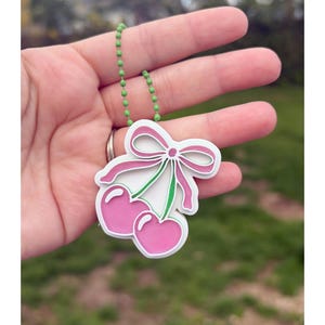 May include: A pink cherry charm with a white outline and a pink bow, connected to a green beaded chain. The charm is held in a hand against a blurred green background.