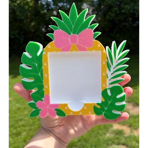 May include: A pineapple-shaped photo frame with a tropical theme. The frame has a yellow and white dotted pattern, green leaves, a pink bow, and a pink flower. The frame is held in a person's hand.