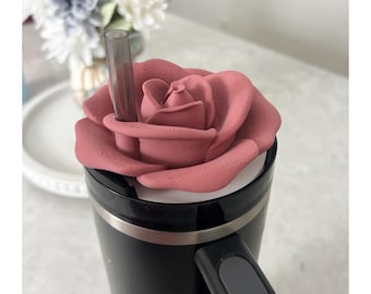May include: A matte dusty rose-colored rose-shaped topper sits atop a black travel mug. The topper features a central opening for a translucent dark gray straw to pass through, with a textured surface mimicking the petals of a blooming rose.