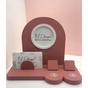 May include: A dusty rose-colored business card holder with a built-in display for a business card and two square holders. The holder has a rounded top with a white circle that has the text "BECdesigns".