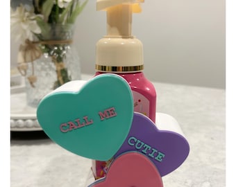 May include: A pink soap dispenser with a beige pump is surrounded by three heart-shaped holders. The holders are teal, purple, and pink, with the phrases "CALL ME", "CUTIE", and "XOXO" embossed on them. The dispenser is on a white tray.