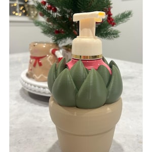 May include: A beige soap dispenser with a green succulent design. The dispenser has a pink top and a beige pump. The base is shaped like a flower pot. A small Christmas tree and gingerbread man are in the background.