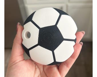 May include: A soccer ball-shaped object in black and white, held in a hand. The object has a hole on the side. The design mimics a classic soccer ball with black pentagons and white hexagons. The object is likely a decorative item or a small toy.