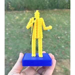 May include: A yellow 3D-printed figure of a person holding a microphone, standing on a blue rectangular base. The figure is made of a spiral-shaped design. The background is blurred green grass and trees.