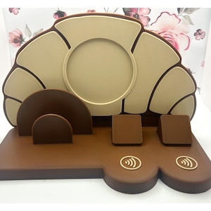 May include: A brown and beige charging station with a croissant-shaped design. It has a circular phone holder, two rectangular charging pads, and a curved support. The base features two wireless charging symbols.