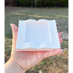 May include: A white and pink book-shaped tray held in a hand. The tray has a rectangular opening in the center, resembling open pages. The edges are detailed to mimic the appearance of a book.