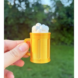 May include: A miniature yellow beer mug with a handle, filled with white foam. The mug has vertical indentations and is held against a blurred green background. The mug is a novelty item.