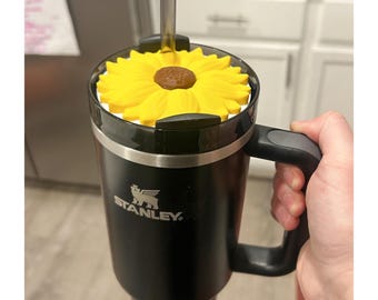 May include: Black Stanley tumbler with a handle, featuring a yellow sunflower design on top. The tumbler has a clear straw and the Stanley logo. The sunflower has a brown center and yellow petals.