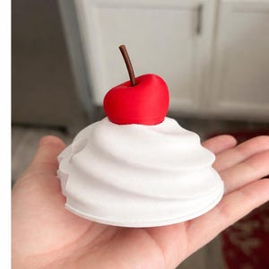 May include: A white, swirled decorative object resembling whipped cream, topped with a bright red cherry with a brown stem. The object is held in a hand, showcasing the detailed design. The overall theme is dessert-inspired.