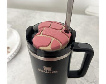 May include: Black Stanley travel mug with a pink and beige lid and a clear straw. The mug has a black handle and the Stanley logo in silver. The lid has a unique, textured design.