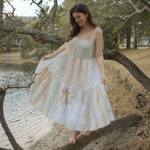 May include: A woman wearing a white and green patchwork dress with a tiered skirt, standing on a tree branch by a lake.