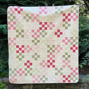 Może przedstawiać: Ręcznie robiona kołdra z kremowym tłem i powtarzającym się wzorem czerwonych i zielonych kwadratów. Kołdra jest wystawiona na zewnątrz, narzucona na kamienny mur, z bujną zielenią w tle.