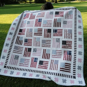 May include: A rectangular quilt featuring a repeating pattern of American flag blocks in red, white, and blue fabric prints. The quilt is bordered by a thin black and white striped frame and an outer edge decorated with small star-shaped motifs.