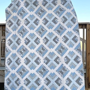 Op de afbeelding: Een blauwe en witte quilt met een geruit patroon. De quilt is gemaakt van vierkanten met een witte achtergrond en blauwe en zwarte geruite vierkanten. De quilt hangt aan een waslijn.