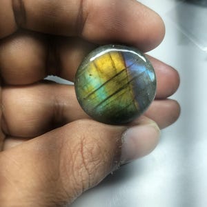 Puede incluir: Una piedra preciosa redonda de labradorita pulida. La piedra muestra colores iridiscentes de amarillo, azul y verde. La gema se sostiene en una mano, mostrando su tamaño y detalle.
