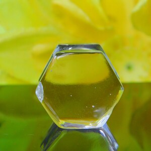 May include: A clear glass hexagonal bottle stopper with a yellow liquid inside. The bottle stopper is sitting on a reflective surface.