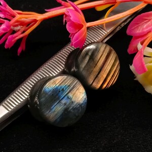 May include: Two circular labradorite ear plugs with iridescent blue and gold flashes, positioned next to a metal ruler showing measurements in inches. The stones feature a polished surface with natural grey and dark inclusions.