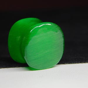 May include: A vibrant green ear plug with a smooth, polished surface. The plug has a rounded, cylindrical shape with a flat, circular face. The color is a bright, saturated green, and the material appears to be glossy.