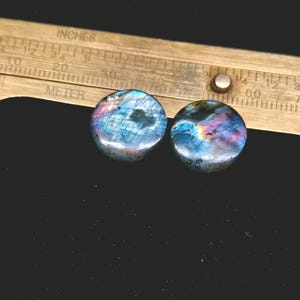 May include: Two round labradorite cabochons featuring iridescent blue, purple, and gold flashes. The stones are positioned below a brass ruler showing a diameter of approximately 0.5 inches for each piece, with visible markings for inches and metric units.