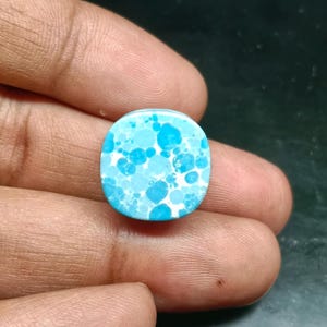 May include: A small, rounded square cabochon featuring a vibrant pattern of various shades of blue and turquoise circular spots set against a bright white background. The surface appears smooth and polished, displaying a distinct speckled aesthetic.
