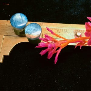 May include: Two circular blue and grey labradorite ear plugs resting on a brass caliper. The caliper shows a measurement of approximately 0.75 inches. A small branch with bright pink flowers is positioned across the metal tool.