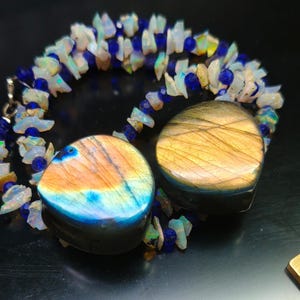 Peut inclure: Un collier orné de deux grandes pierres de labradorite polies, aux couleurs irisées de bleu, orange et or. Le collier est agrémenté d'un fil de petites opales irrégulières et de perles bleues, créant un bijou unique.