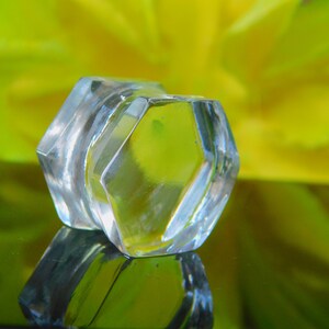 May include: A clear glass bottle stopper with a hexagonal shape. The stopper is sitting on a green surface with a yellow flower in the background.