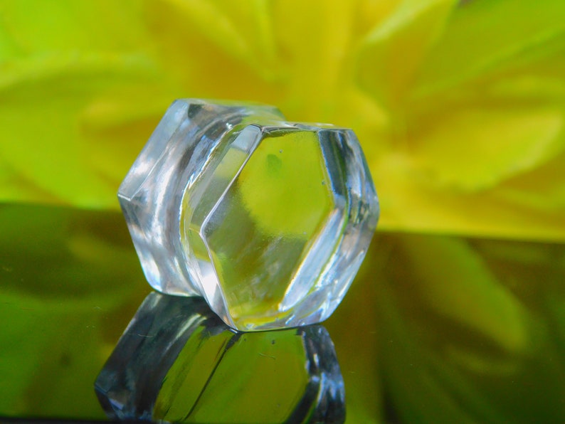May include: A clear glass bottle stopper with a hexagonal shape. The stopper is sitting on a green surface with a yellow flower in the background.