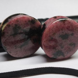 May include: Two round, polished plugs with a pink and black mottled pattern. The plugs are made of stone and are set against a blurred background with a black object and a black cord.