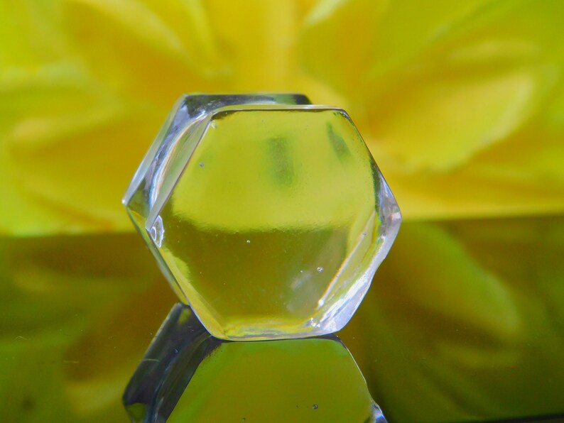 May include: A clear, hexagonal glass ice cube sits on a yellow reflective surface.