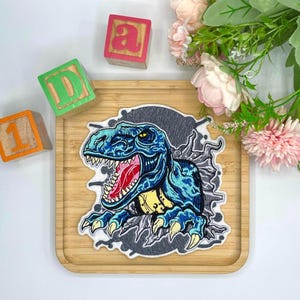 Könnte beinhalten: Ein blau-schwarzer Dinosaurier-Aufnäher mit rotem Maul und scharfen Zähnen. Der Aufnäher liegt auf einem Holztablett mit bunten Holzblöcken und Blumen im Hintergrund. Der Dinosaurier bricht durch einen grauen Hintergrund.