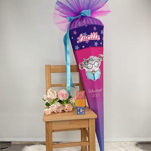 Könnte beinhalten: Eine große, kegelförmige Schultüte in Rosa und Lila, verziert mit einer Cartoon-Illustration eines lesenden Kindes. Die Tüte ist mit dem Namen "Lucille" und dem Text "Schulkind 2024" personalisiert. Sie ist mit einer Tüllschleife versehen.