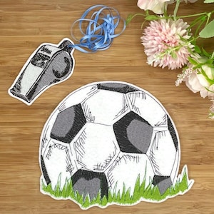 Könnte beinhalten: Gestickte Fußball- und Pfeifenaufnäher. Der Fußball ist weiß mit schwarzen Fünfecken, eingebettet in grünes Gras. Eine grau-weiße Pfeife mit blauem Band ist ebenfalls vorhanden. Die Aufnäher befinden sich auf einer Holzoberfläche.