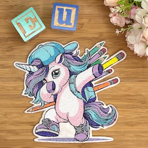 Könnte beinhalten: Gestickter Aufnäher mit einem Einhorn, das dabbt, eine blaue Kappe und Turnschuhe trägt, mit einem Rucksack und Bleistiften. Das Einhorn hat lila und blaues Haar vor einem Holzgrund. Zwei Holzblöcke mit Buchstaben befinden sich im Hintergrund.