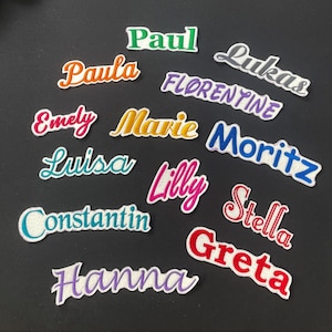 K&ouml;nnte beinhalten: Eine Sammlung gestickter Namensaufn&auml;her in verschiedenen Farben und Schriftarten, darunter "Paul", "Paula", "Emely", "Marie", "Luisa", "Lilly", "Constantin", "Hanna", "Lukas", "Florentine", "Moritz", "Stella" und "Greta". Jeder Aufn&auml;her hat einen wei&szlig;en Rand.