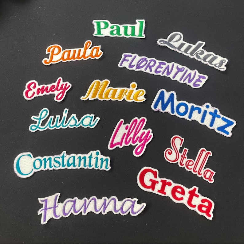 K&ouml;nnte beinhalten: Eine Sammlung gestickter Namensaufn&auml;her in verschiedenen Farben und Schriftarten, darunter "Paul", "Paula", "Emely", "Marie", "Luisa", "Lilly", "Constantin", "Hanna", "Lukas", "Florentine", "Moritz", "Stella" und "Greta". Jeder Aufn&auml;her hat einen wei&szlig;en Rand.