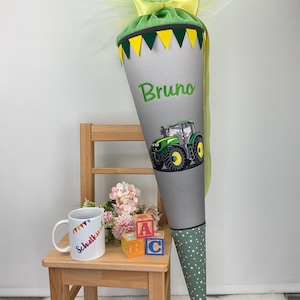 Puede incluir: Un cono escolar gris y verde con un dise&ntilde;o de tractor y el nombre "Bruno". El cono est&aacute; decorado con detalles amarillos y verdes, una parte superior de tul verde y una base de lunares verdes y blancos.