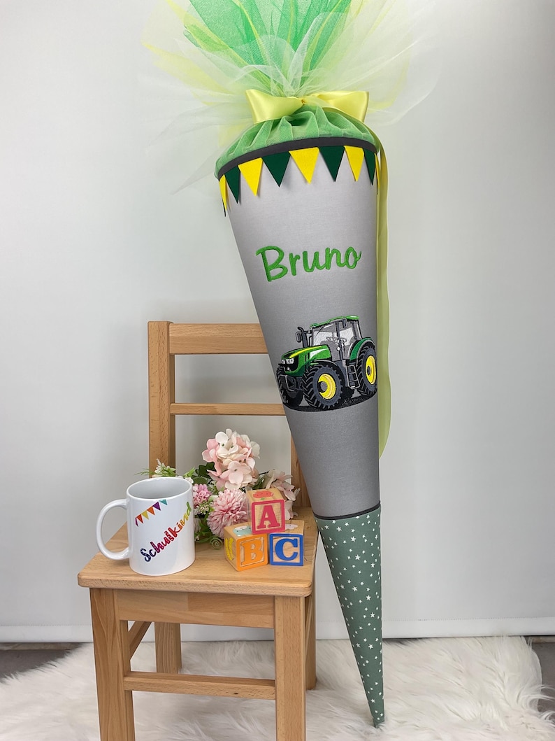 Puede incluir: Un cono escolar gris y verde con un dise&ntilde;o de tractor y el nombre "Bruno". El cono est&aacute; decorado con detalles amarillos y verdes, una parte superior de tul verde y una base de lunares verdes y blancos.