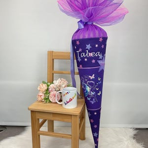Könnte beinhalten: Eine lila Schultüte mit dem Namen "Tabea" in weißer Schrift, verziert mit Sternen und Schmetterlingen. Die Tüte ist mit Lagen aus lila und rosa Tüll und einem passenden Band versehen. Ein kleiner Holzstuhl hält einen Becher mit dem Text "Schulkind".