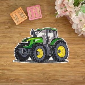 Könnte beinhalten: Gestickter Aufnäher eines grünen und schwarzen Traktors mit gelben Rädern. Der Traktor hat eine weiße Umrandung und befindet sich auf einem Holzgrund. Zwei Holzblöcke mit den Buchstaben "T" und "R" sind zu sehen.