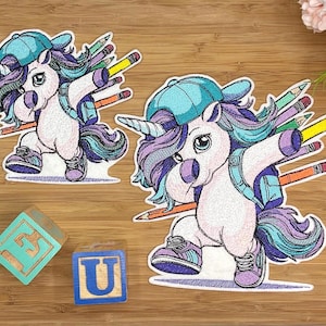 Könnte beinhalten: Zwei gestickte Aufnäher mit einem weißen Einhorn mit violetter und türkisfarbener Mähne, das eine Baseballkappe, einen Rucksack und violette Turnschuhe trägt. Das Einhorn macht einen Dab, umgeben von bunten Stiften. Zwei Holzwürfel zeigen E und U.