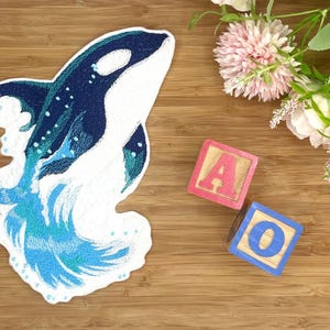 Könnte beinhalten: Ein gestickter Aufnäher mit einem stilisierten Orca, der aus dem Wasser springt. Der Orca ist hauptsächlich weiß und dunkelblau, mit türkisfarbenen Akzenten. Zwei Holzblöcke mit den Buchstaben "A" und "O" sind ebenfalls vorhanden, zusammen mit rosa Blumen.