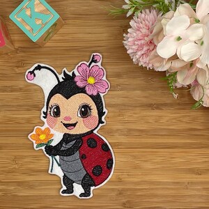 Könnte beinhalten: Gestickter Aufnäher einer Cartoon-Marienkäferin, die eine Blume hält, mit einem rot-schwarz gepunkteten Panzer, einem schwarzen Kopf und einer rosa Blume auf dem Kopf. Der Aufnäher befindet sich auf einer Holzoberfläche mit Spielzeugblöcken und Blumen im Hintergrund.