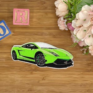 Könnte beinhalten: Grüne Lamborghini Sportwagen Stickerei. Der Patch befindet sich auf einer Holzoberfläche mit rosa und blauen Buchstabenblöcken.